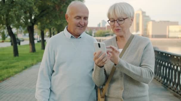 Långsam rörelse av äldre man och kvinna chatta skrattar tittar på smartphone skärm promenader i staden — Stockvideo