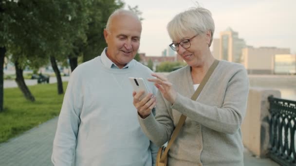 Slow motion van senior paar lopen langs de rivier dijk praten met behulp van smart phone op zomerdag — Stockvideo
