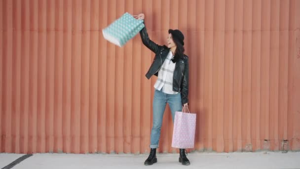 Portret van een vrolijke jongedame die boodschappentassen optilt en buiten lacht tegen de achtergrond van de muur — Stockvideo