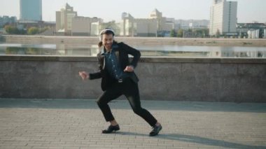 Kulaklıkla müzik dinleyip nehir kenarında dans eden genç ve mutlu bir işadamı.