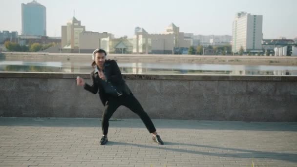 Fröhlicher Typ mit Kopfhörern tanzt draußen am Flussufer und genießt Musik — Stockvideo