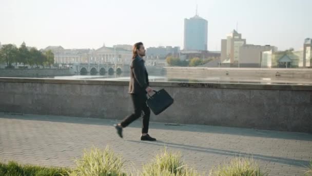 一个野心勃勃的年轻商人提着公文包沿着河堤走在城市里 — 图库视频影像