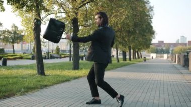 Takım elbiseli neşeli işadamının şehir sokaklarında mutluluğu ifade eden evrak çantasıyla eğlenen bir portresi.