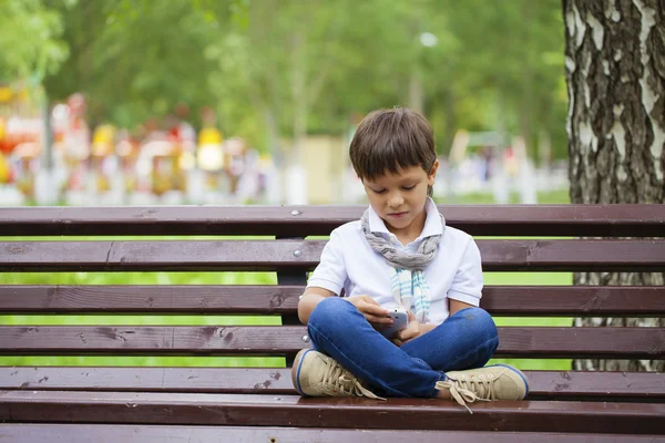 Een kleine jongen zit op een bankje en bellen door mobiele telefoon — Stockfoto