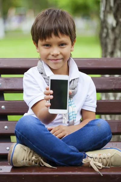 Malý chlapec sedí na lavičce a zobrazení displeje mobilního telefonu — Stock fotografie