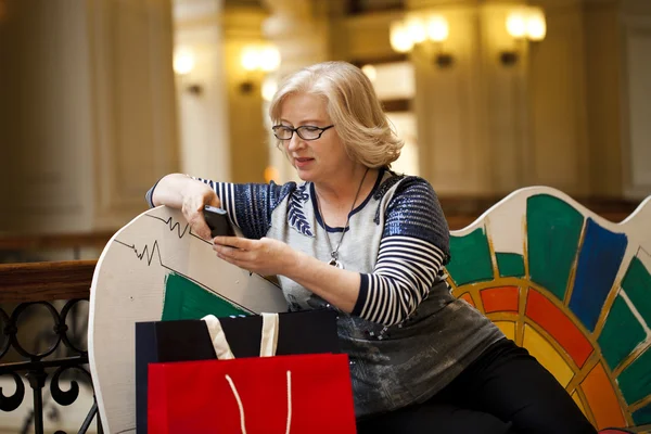 Gammal blond kvinna ringer via telefon med kassar och påsar — Stockfoto