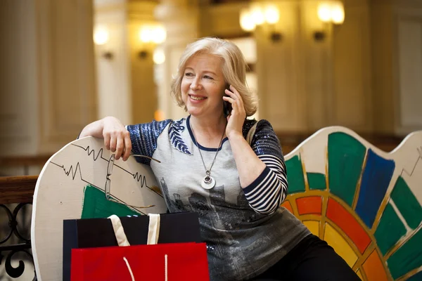 Gammal blond kvinna ringer via telefon med kassar och påsar — Stockfoto