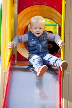 Blonde little boy sits on a childrens slide at the playground clipart
