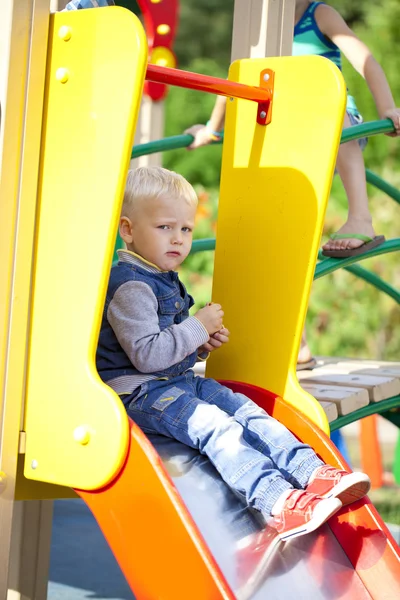 Blonde little boy sits on a childrens slide at the playground — Stock Photo, Image