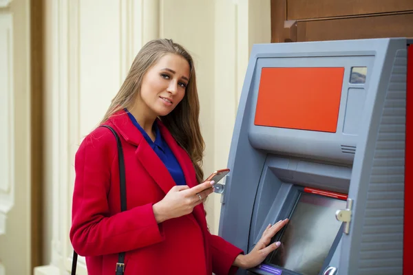 Young blonde woman in a red coat withdraw cash from an ATM in th — Stock Photo, Image