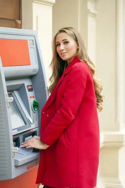 Mujer rubia joven en un abrigo rojo retirar dinero en efectivo de un cajero automático en th — Foto de Stock