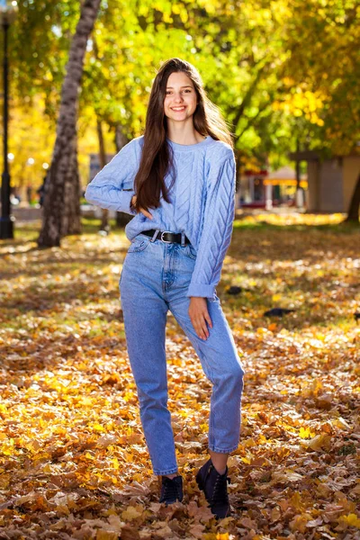 秋の公園でポーズをとるウールのセーターと青いジーンズの若い女の子の完全な長さの肖像画 — ストック写真