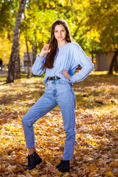 秋の公園でポーズをとるウールのセーターと青いジーンズの若い女の子の完全な長さの肖像画 — ストック写真