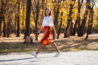 Sonbahar parkında yürüyen turuncu etekli esmer bir kadının vücut portresi.