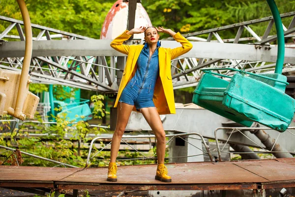 Bir lunaparkta atlıkarınca arka planında poz veren sarı ceketli ve kot pantolonlu manken.