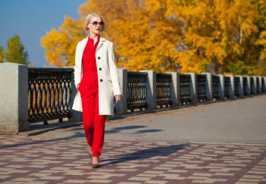 Güz parkında bej ceketli güzel sarışın kadın yürüyor.