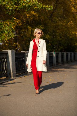 Güz parkında bej ceketli güzel sarışın kadın yürüyor.