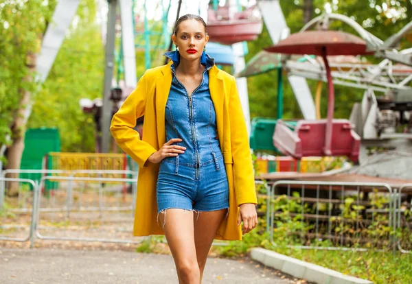 Модна Модель Жовтому Пальто Джинсовий Комбінезон Позує Карусельному Фоні Парку — стокове фото