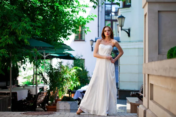 Beyaz elbiseli, yaz sokağı pozu veren güzel bir kadının uzun bir portresi.