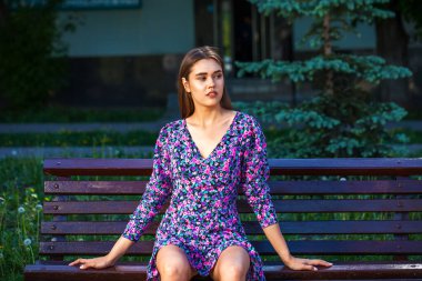Parktaki bankta otururken poz veren leylak rengi bir yaz elbisesi içindeki güzel kız.