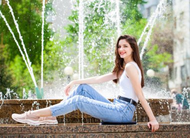 Bir şehir çeşmesinin arka planında poz veren güzel bir kızın tam boy portresi.