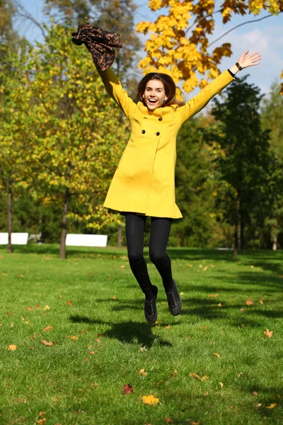 Happy woman in yellow coat jumping in autumn park — Stock Photo, Image