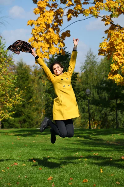 Happy woman in yellow coat jumping in autumn park — Stock Photo, Image