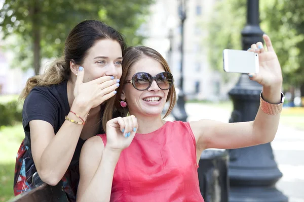 Chicas jóvenes sonrientes con teléfono celular sentadas en un banco en un parque —  Fotos de Stock