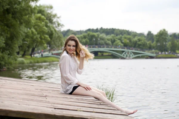 Junge schöne Mädchen in einer weißen Tunika sitzt auf einem hölzernen Pier auf t — Stockfoto