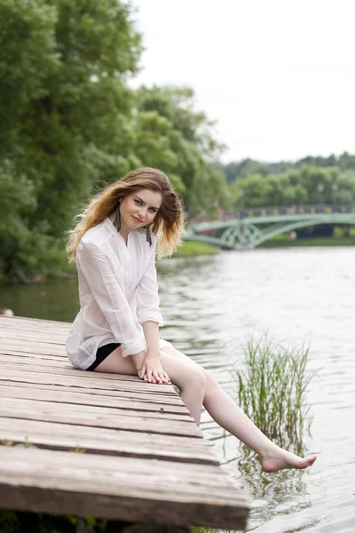 Giovane bella ragazza in una tunica bianca si siede su un molo di legno su t — Foto Stock