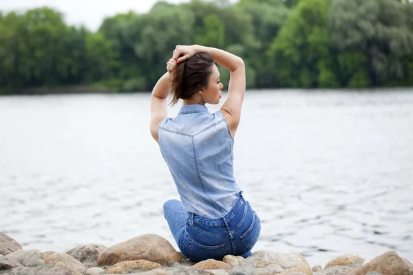 年轻漂亮的黑发女孩坐在河上的岩石上 — 图库照片