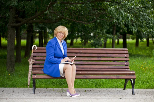 Oudere zakenvrouw in jas sittin op Bank met dagelijks een logboek bij — Stockfoto