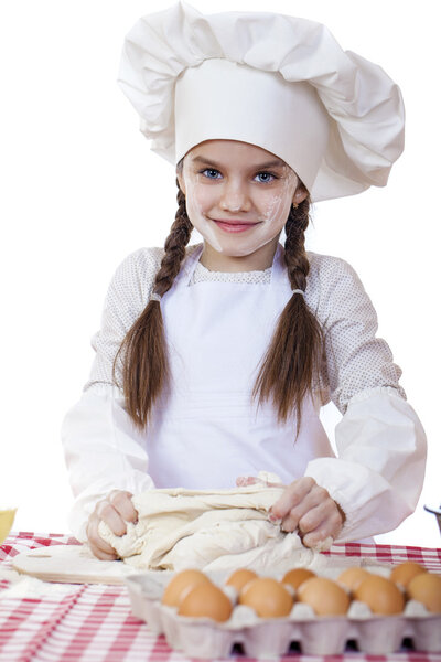 Little girl in a white apron and chefs hat knead the dough in th