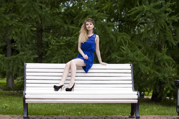 Menina loira bonita em vestido azul sentado em um banco no verão — Fotografia de Stock