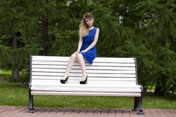 Красивая блондинка в синем платье сидит на скамейке летом — стоковое фото