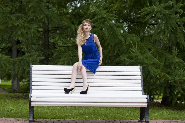 Menina loira bonita em vestido azul sentado em um banco no verão — Fotografia de Stock