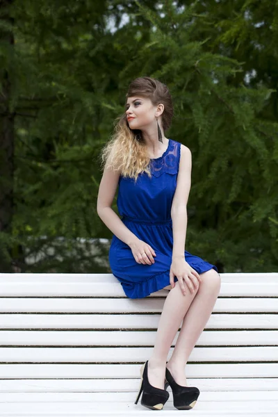 Schöne blonde Mädchen in blauem Kleid sitzt auf einer Bank im Sommer — Stockfoto