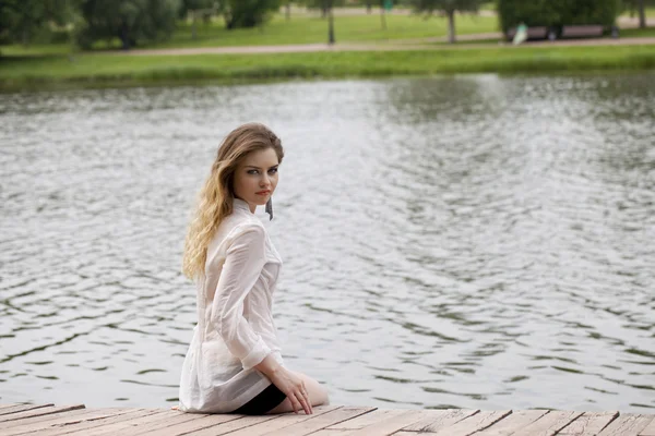 Young beautiful girl in a white tunic sits on a wooden pier on t — Stock Photo, Image