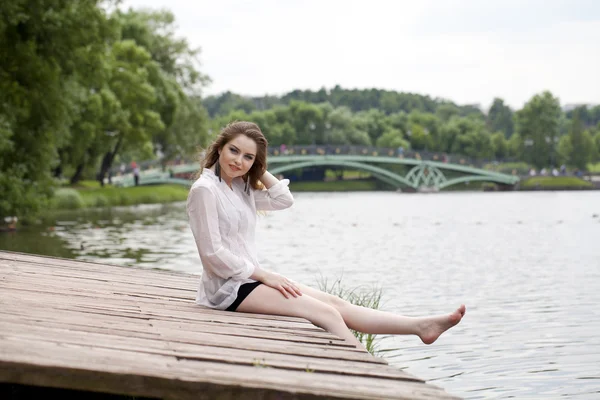 एक सफेद ट्यूनिक में युवा सुंदर लड़की टी पर एक लकड़ी के तट पर बैठती है — स्टॉक फ़ोटो, इमेज