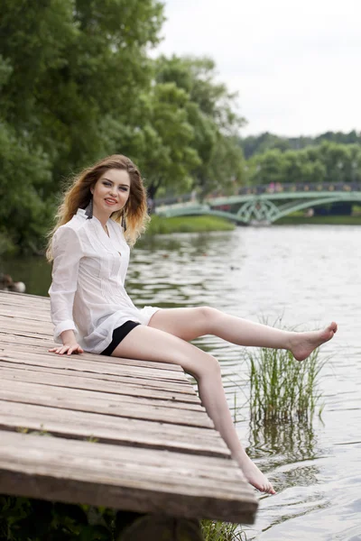 Joven hermosa chica en una túnica blanca se sienta en un muelle de madera en t —  Fotos de Stock