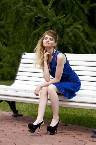 Menina loira bonita em vestido azul sentado em um banco no verão — Fotografia de Stock