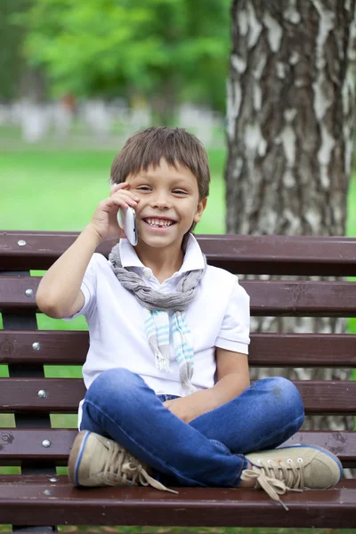 ベンチで携帯電話を呼び出す小さな男の子が座っています。 — ストック写真