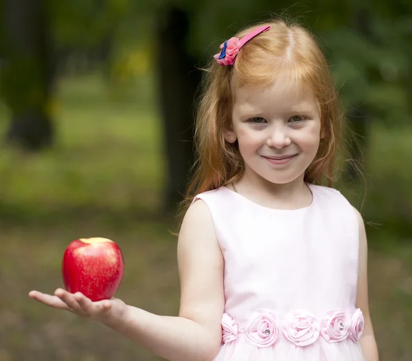 Little red-haired girl holding a red apple on a background of gr — Stock Photo, Image