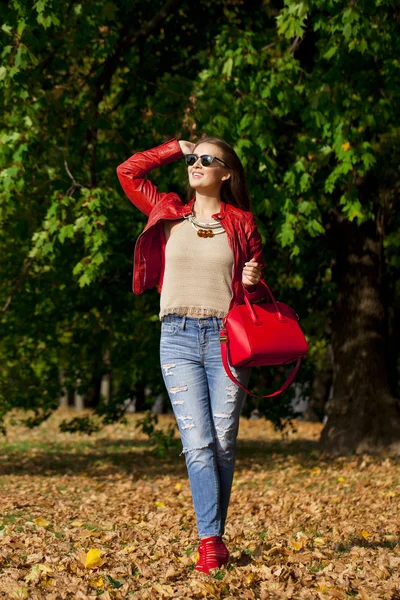 Young woman in fashion red jacket and blue jeans in autumn park — Stock Photo, Image