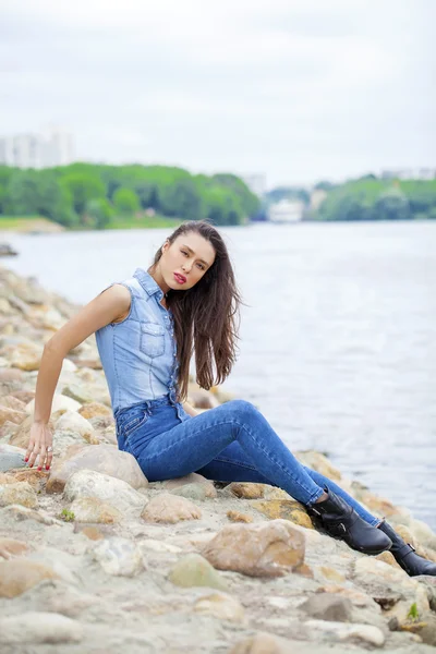 Jonge mooie brunette meisje, zittend op de rotsen op de rivier — Stockfoto