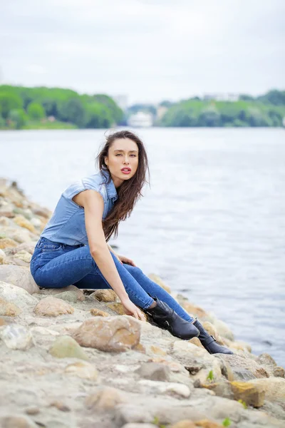 Jonge mooie brunette meisje, zittend op de rotsen op de rivier — Stockfoto