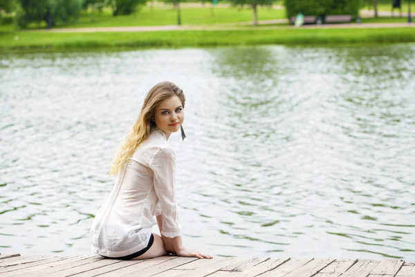 Young beautiful girl in a white tunic sits on a wooden pier on t — Stock Photo, Image