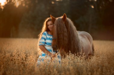 Beautiful long-haired young woman with red tinker horse in oats field at sunset