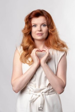 Genç kızıl saçlı kadın, günlük kıyafetler ve gözlük takıyor, gülümsüyor, elleriyle kalp sembolü çiziyor..