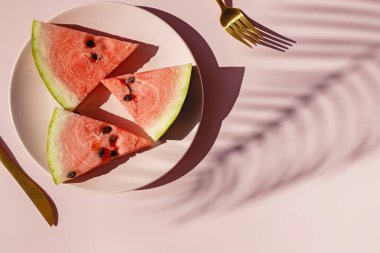 Pembe tabakta taze kırmızı karpuz dilimleri ve tropikal yaprak gölgesi.
